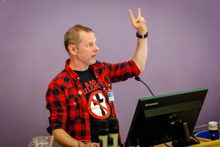 The author, a straight sized white man with short grey hair, standing behind a screen and microphone wearing a bad religion t shirt. 