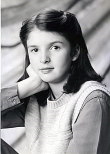 Emily, a nine year old white girl with shoulder length brown hair pinned on either side away from her face. She is leaning her head on her hand and is wearing a sweater vest over a blouse. The photo is in black and white. 