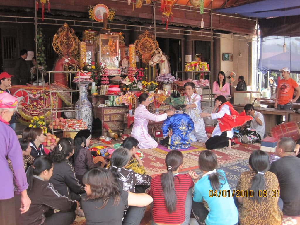 A len dong ceremony. Four people wearing traditional outfits sit in front of an alter on a rug surrounded by an audience who are sitting on the floor around them. 