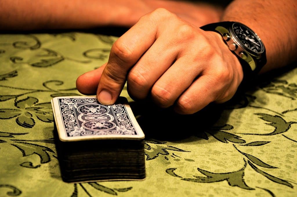 A white person's hand with a face-down blue and white deck of cards on a patterned tablecloth. In the photo, the person is sitting opposite the viewer, with their index finger and thumb resting on the top card.