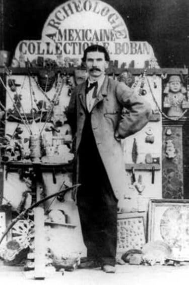 A black and white photograph of a white, slender man with dark hair and a dark moustache. He is wearing a suit and a long coat and standing in front of a board of artifacts. 