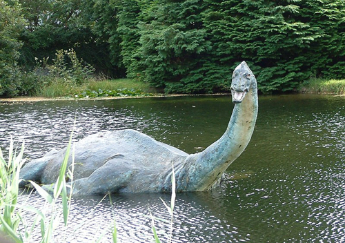 A coloured statue of "Nessie" the sea monster in a lake at the Nessie Museum, with green trees behind. She looks like a plesiosaur (ancient and extinct aquatic reptile).
