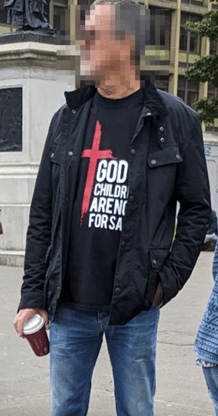 A man wearing a "God's children are not for sale" t shirt with a black jacket over the top. His face has been pixelated. 

Source: Brian Eggo