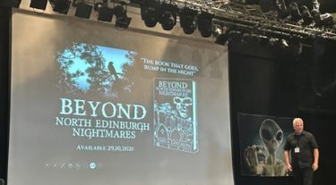 A white man with grey hair wearing a lanyard stands to the right of a stage, with an image of a stereotypical alien behind him, and a slide projected to his right advertising a book called "Beyond", North Edinburgh Nightmares.
Source: Brian Eggo