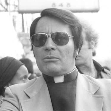 A black and white photograph of Jim Jones wearing a clerical collar and dark sunglasses. 

Nancy Wong, CC BY-SA 4.0 <https://creativecommons.org/licenses/by-sa/4.0>, via Wikimedia Commons
