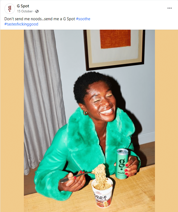 A screenshot of a facebook post by G Spot. Text says "Don't send me noods...send me a G Spot #soothe #tastesfxckinggood" and accompanies a photo of a grinning black woman with her eyes closed, wearing a bright green, fluffy collar-and-cuffs leather jacket, holding a green can of G Spot soothe and eating a beef ramen noodle cup. She has red painted nails and is sitting at a wooden table.