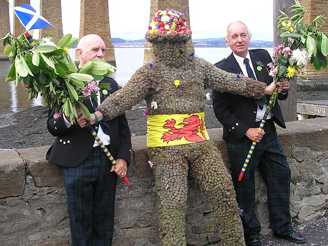Two men in uniform flank a central 'Burry Man', a Scottish traditional dress in which a man is covered in burrs, hat covered in flowers and a sash made of a folded Royal Standard of Scotland, showing the red dragon on a yellow background. He holds two  waist-high poles decorated withh leaves and flowers (to support himself as he walks). Image by Michael Mabbott.
