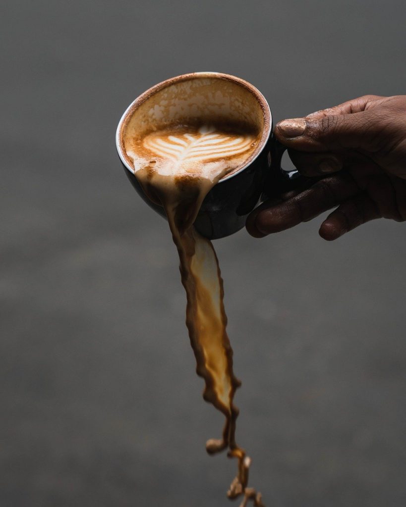 A person with dark skin tips and spills coffee from a cup. The image freezes the liquid in time, with coffee tumbling down through the air over a dark grey background towards the floor. Image by poedynchuk from Pixabay