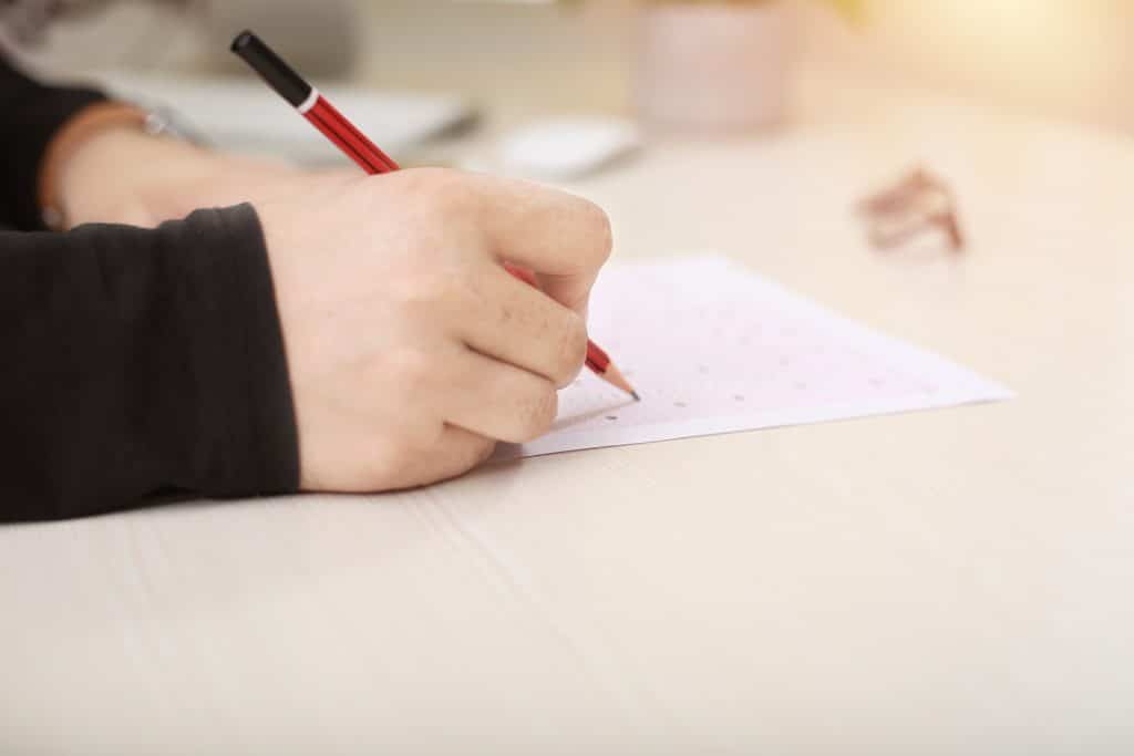 View of a test paper with someone's hand writing on it - a white student holds a red and black pencil in their right hand and is writing on the test paper on a cream-coloured desk