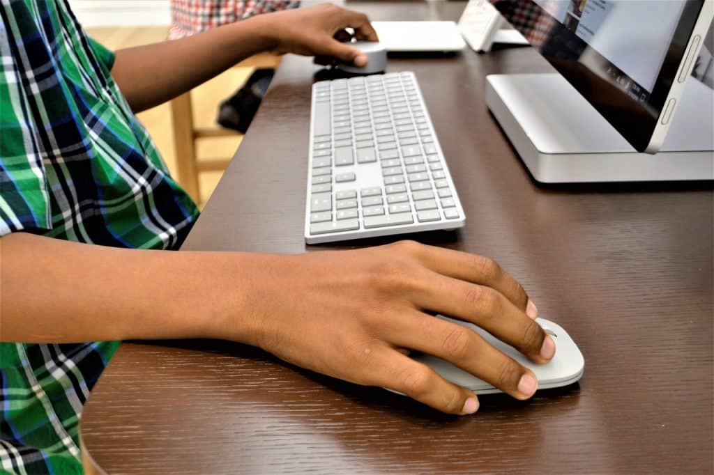 A young man sits at a desk using a computer via a monitor, with separate wireless mouse and keyboard.