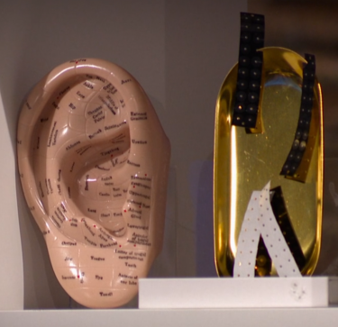 A screenshot of the BBC Dragon's Den episode, showing a close-up of a ceramic model of an ear acupuncture map, including specific application points with corresponding body parts. Next to it is a golden tray with packs of ear seeds on display.