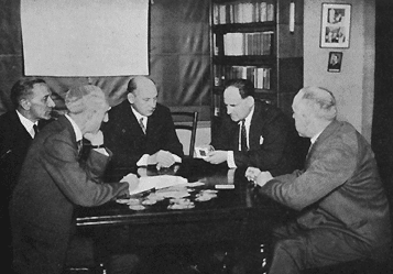 A black and white photograph of five white men, all in suits and appearing to be over ~50 years of age. They are sat around a table that has cards spread out on it, with a projector screen and bookcase behind them.
