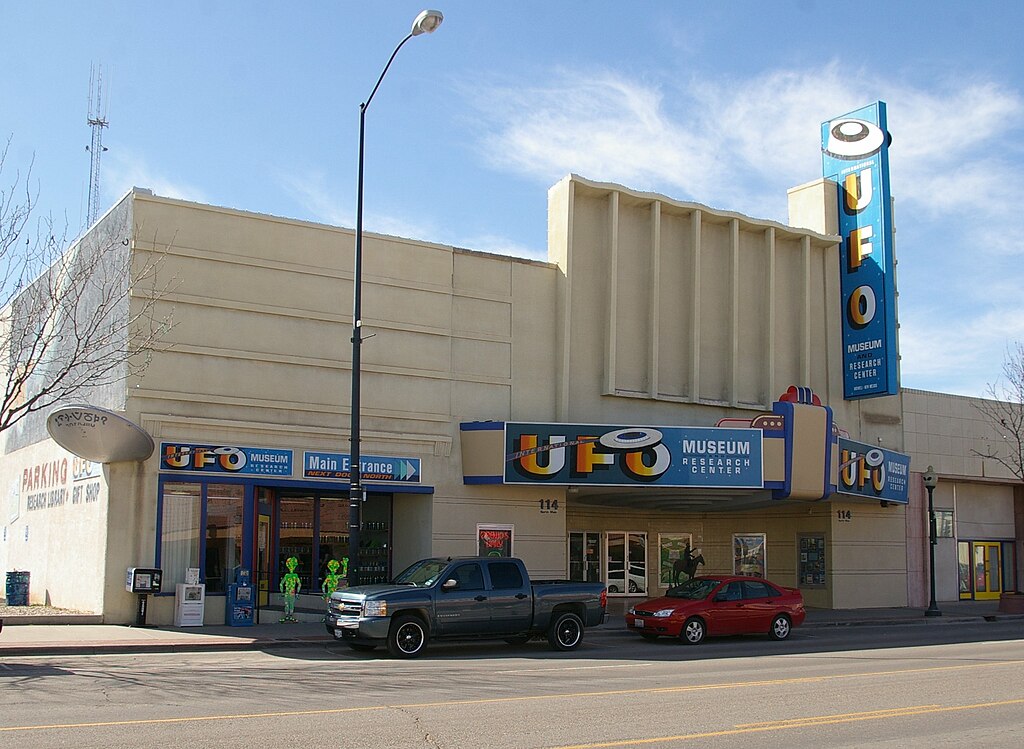 Photograph of the Roswell UFO Museum in New Mexico showing the front exterior. A truck and a car are parked outside and four different signs say UFO Museum Research Centre. Two green alien figures stand in front of its windows.