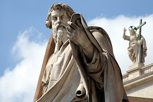 A statue of St Paul (sculptor, Adamo Tadolini), he looks down and towards the camera, wearing robes and holding up his left arm. He has a long beard. In front of the Basilica in the Vatican, a similar statue of Christ is visible behind him to the right of the image.