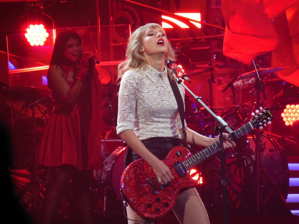 Taylor Swift singing on stage, in red light and playing a red guitar.