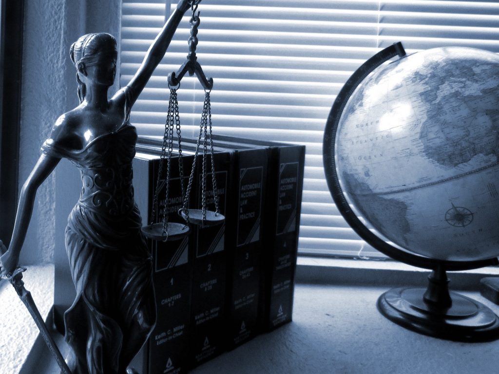 Monochrome photo of a statuette of Lady Justice holding her sword and scales, blindfolded, by some books and a globe on a desk