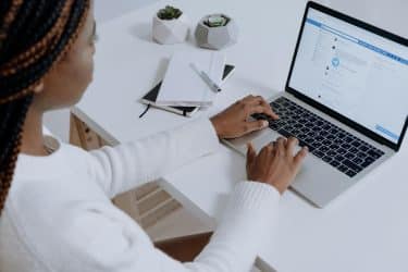 A Black woman in a long-sleeved white top sitting at a white desk browses Facebook on her laptop