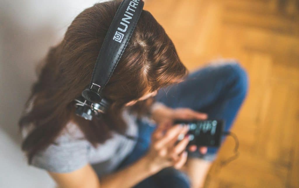 A woman with long brown hair sits cross-legged with black over-ear headphones on, which are plugged into her smartphone, as she uses one hand to navigate its screen