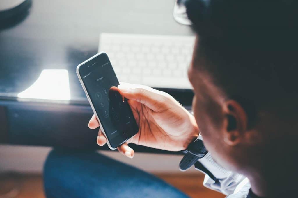 A man with short dark hair and light brown skin looks at his smartphone as he unlocks it with his right hand, his bright white keyboard out of focus in the background