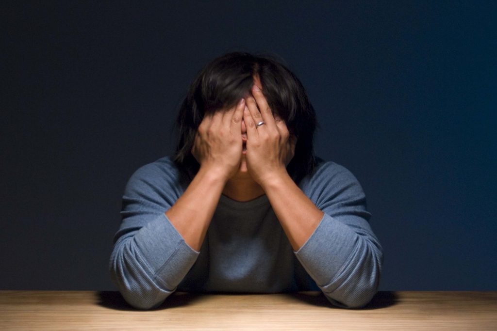 A person in a blue jumper with pushed-up sleeves sits behind a wooden surface with their elbows resting on it and their face covered behind their hands. They have dark, shoulder-length, shiny hair that covers some of their fingertips as it falls forward