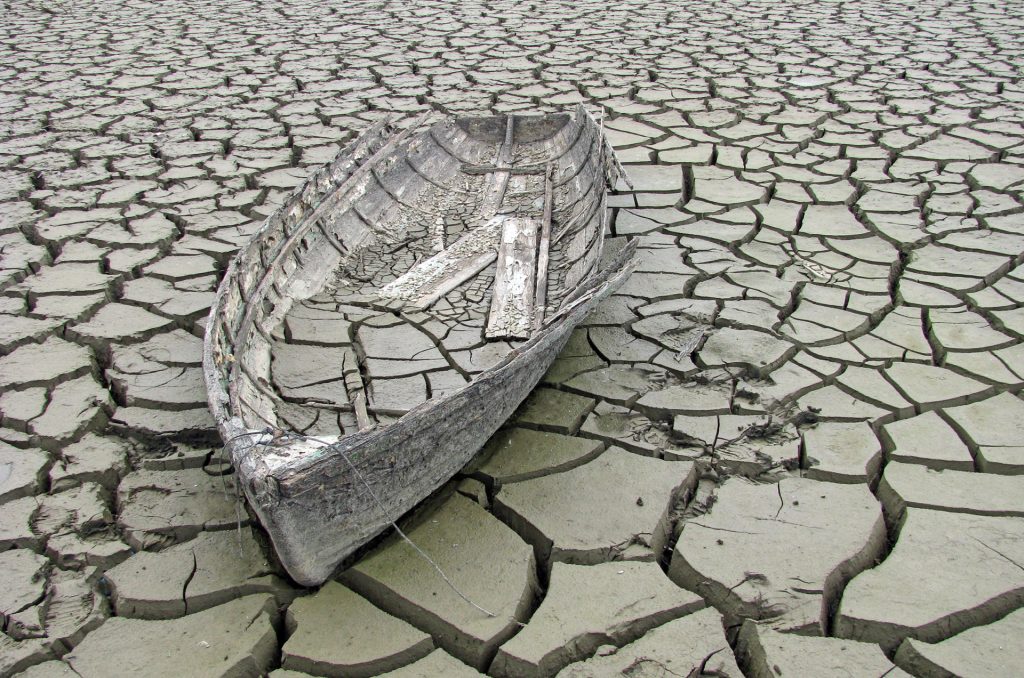 An old wooden rowboat sits atop a large, cracked mud flat, with some of the same cracked pattern inside the boat where mud has flooded in and dried out. The whole scene has a sepia tone but the photod oesn't seem to be false coloured.