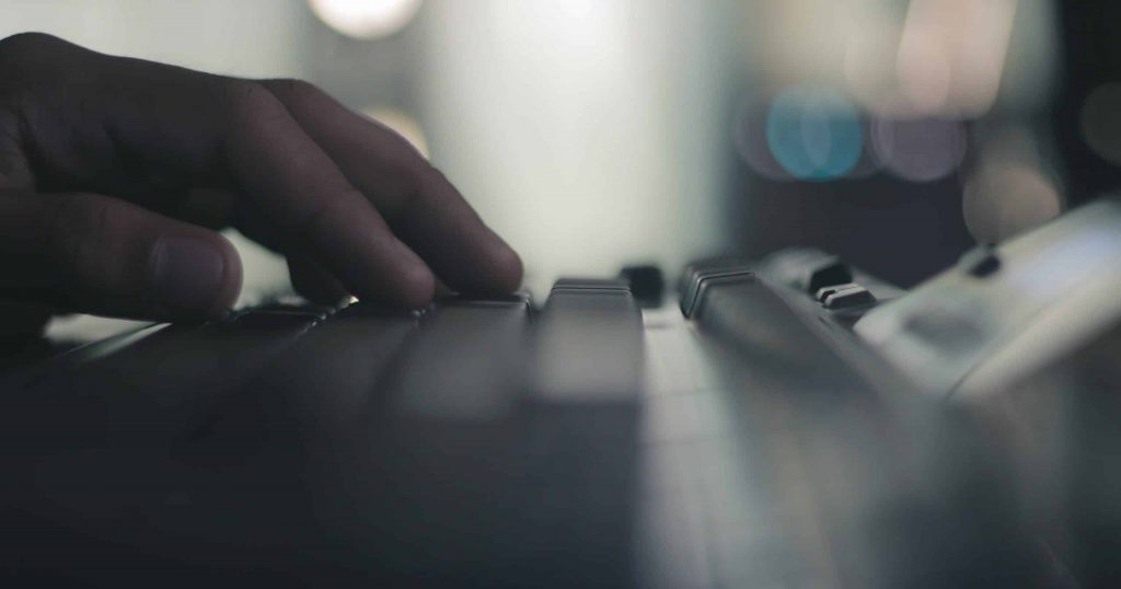 A hand typing on a computer keyboard in a dimly lit room, the fore and background out of focus