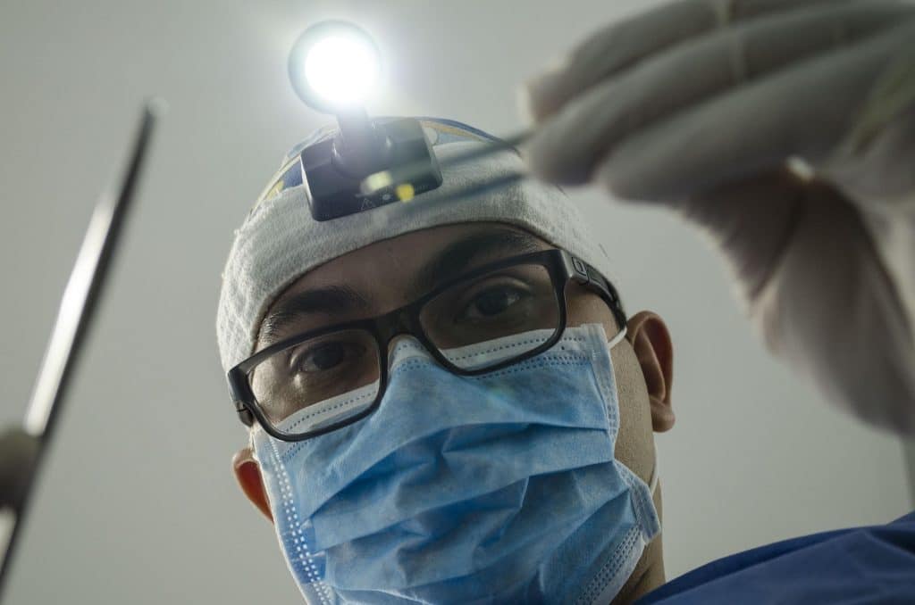 A dentist looms over someone's face/the camera, holding a metal implement in each hand, with a headtorch shining down from his forehead. He's wearing glasses whose rims are resting on a blue surgical mask, and he's wearing beige gloves.