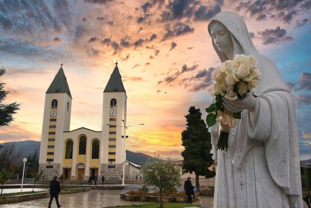 Against a dramatic, colourful sky, a statue of the Virgin Mary holds a bouquet of cream roses and a rosary chain outside the double-steepled church at Medjugorje. Rolling mountains in the background contrast with a wide, wet paved ara in the foreground