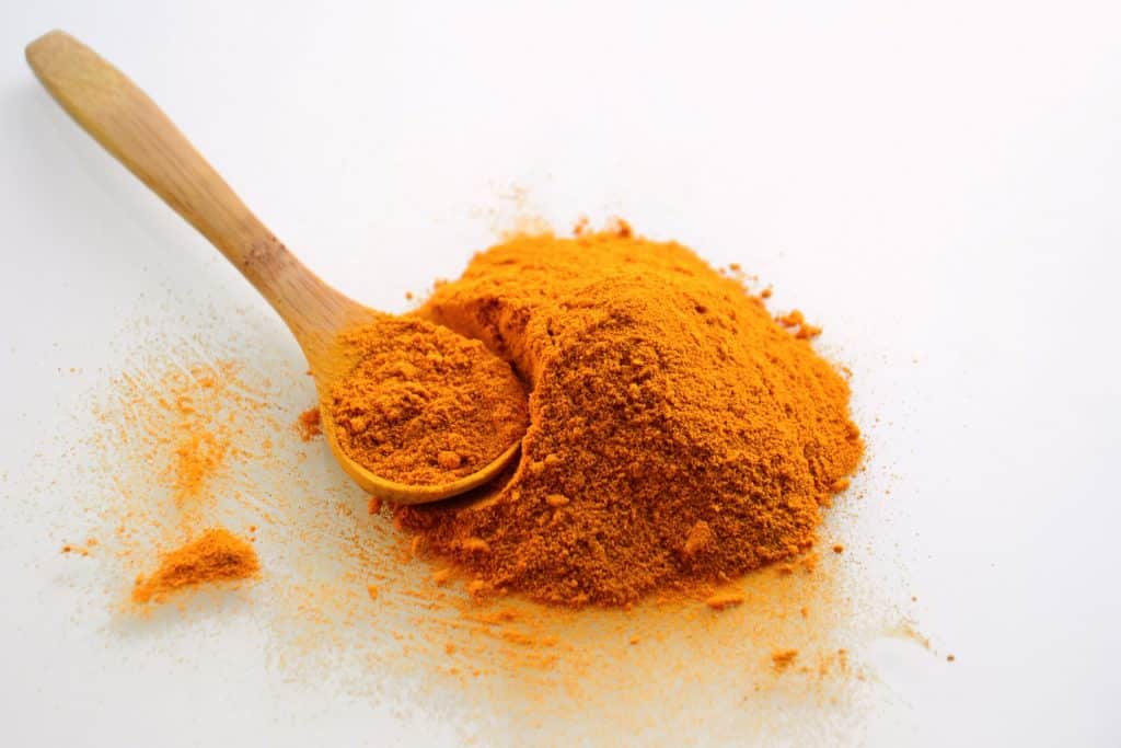 A pile of turmeric powder on a white surface, with a wooden spoon holding some of the powder to the left, resting beside the pile.