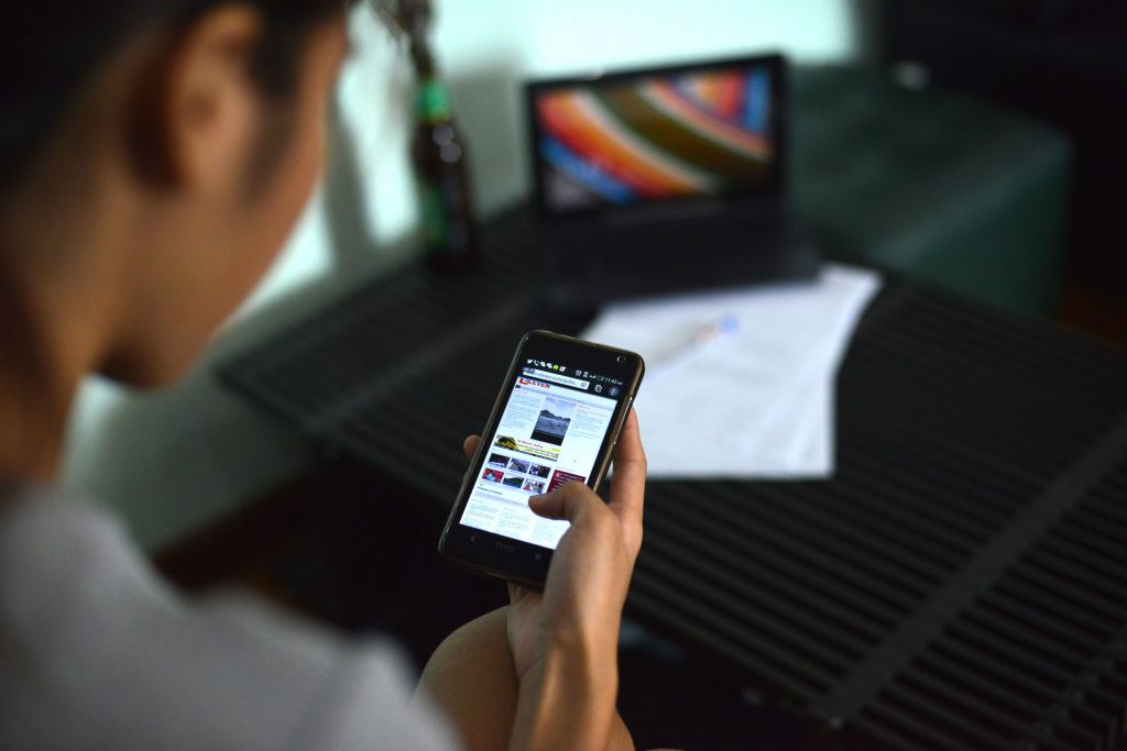 A woman browses the internet on her mobile phone, with her laptop, a small glass bottle (like many beer brands) and some papers out of focus on a desk below