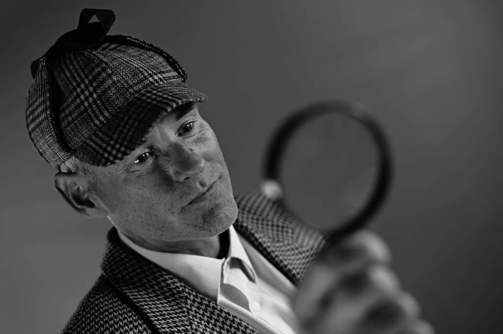 A man dressed in the Sherlock Holmes-esque deerstalker hat and a suit with patterned jacket holds up a magnifying glass in front of him. The photo is in black and white and taken at an angle