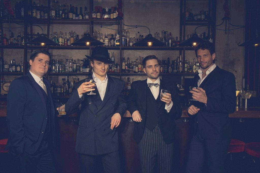 Four men stand, leaning on the bar behind them. Behind the bar itself are shelves covered in drink bottles, and some low-hanging, low-light lampshades are above it. The men are dressed as if they're wealthy, in suits - old money-style outfits including bow tie, long pinstripe jackets, waistcoats, all in dark colours.