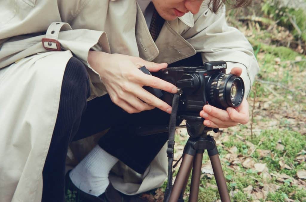 A female-presenting white person crouches on grassy and leafy ground while adjusting a DSLR camera on a tripod. She's wearing a tan trenchcoat, white socks, dark trousers and dark shoes
