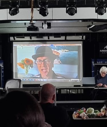 The conference stage. There is a pre-recorded video of a man in a Bowler Hat on the screen, presumably Mark Byford.