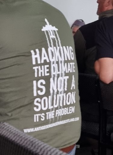 The back of a man in a khaki green t-shirt which says "Hacking the climate is not a solution, it's the problem" with an image of a plane.
