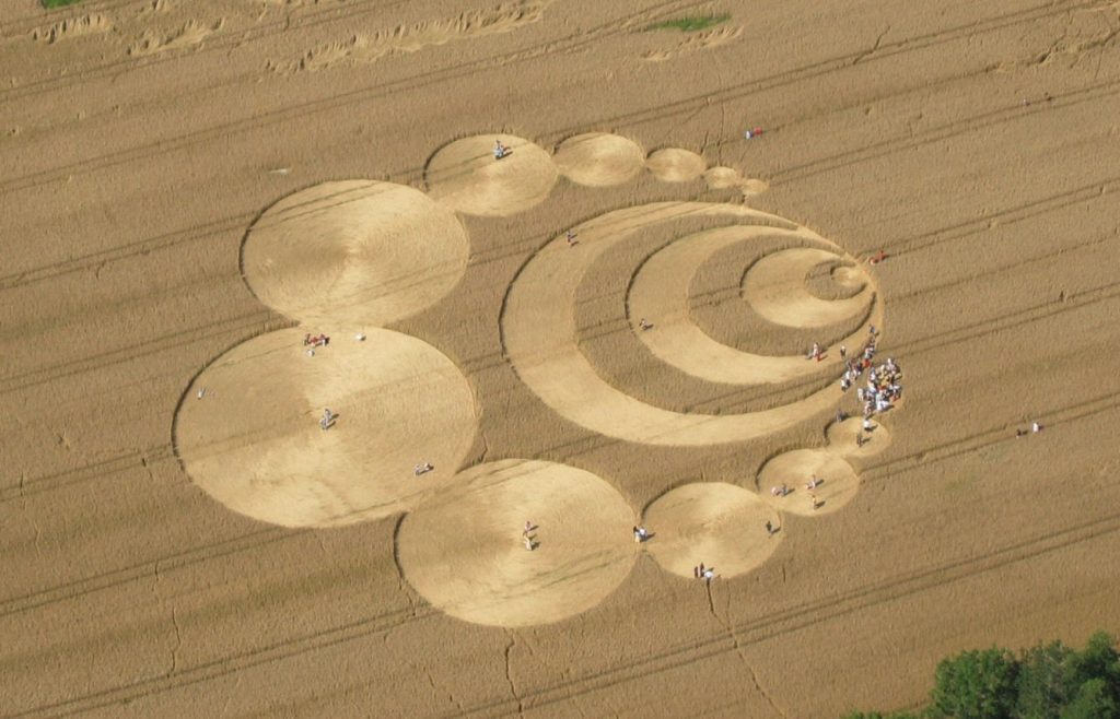 An aerial photo of an area of crop circles in a wheat field in Switzerland. The circles begin small to the right of the image, radiating outwards, and surrounded by an open ring of circles increasing in size towards the curve's apex. People are walking the flattened stem areas.