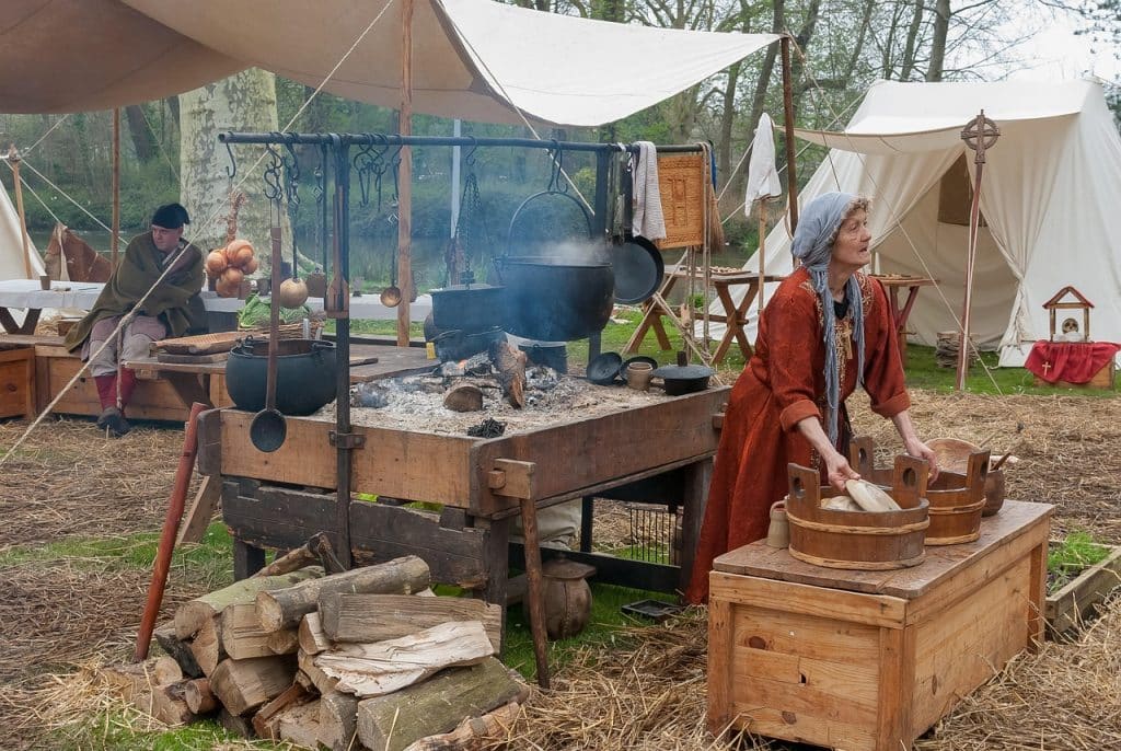 A white woman in a red dress and blue headscarf arranges (anachronistic) ceramic plates in a wooden bowl at a medieval reenactment site in the woods, with small black steaming/smoking hanging cauldrons or pans behind her, under a canvas shelter. A man sits in the background. There's a pile of chopped wood on the grass and some Christian symbols in the scene, with a second, more elaborate tent in the background.