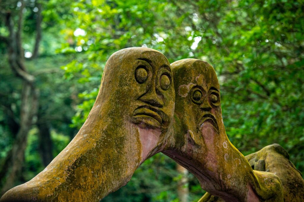 The sculpture shows two human faces carved at the apex of a stone arch, in a forest grove.