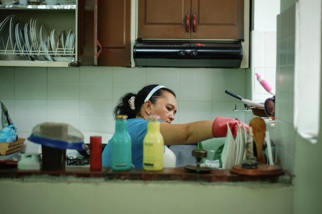 A woman wearing a white headband and red cleaning gloves, with her hair in a ponytail, moves items in the kitchen. Only her upper body is visible. She's by an extractor fan and there's an open cupboard with a rack of plates inside. The decor is quite dated.