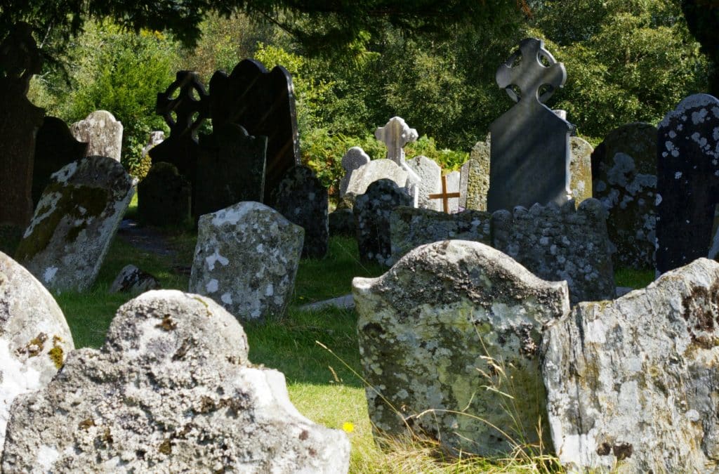 Photo of a partly sunny and partly shaded grassy graveyard with old, worn stones covered in lichens. Some crosses are visible.