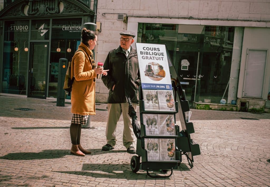 Jehovah's Witnesses in France offering bible courses. A man in a flat cap speaks with a woman in a mustard coat on the street, both standing behind the stand holding copies of the religion's literature.