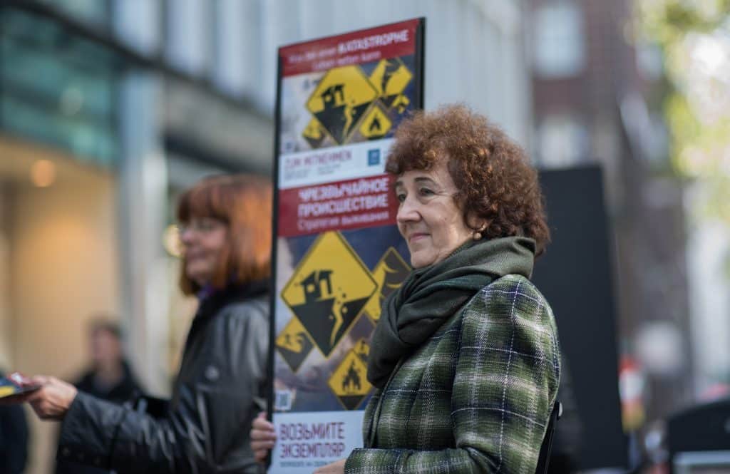 Jehovah's Witnesses in Germany warning about End Times. A woman holds a sign with alarming red and yellow graphics and Russian text. Another behind her hands out literature.