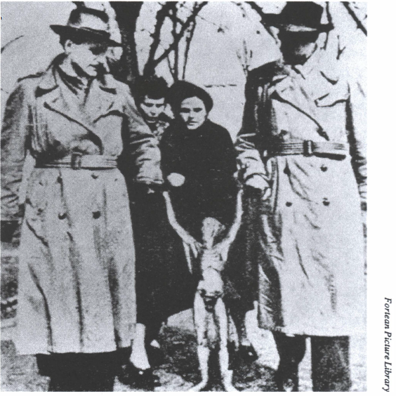 The Silver Man 'alien' photo, a low-quality black and white image showing two men in trenchcoats and wide-brimmed hats holding the 'hands' of a waist-height 'silver man' alien figure, with two women in the background observing (from the Fortean Picture Library)