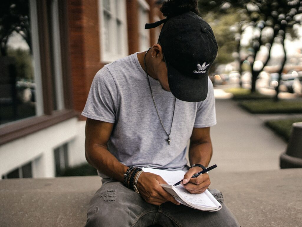 A man sits on a wall in the street, wearing a black Adidas cap, necklace and grey t-shirt, writing in an A4 notepad