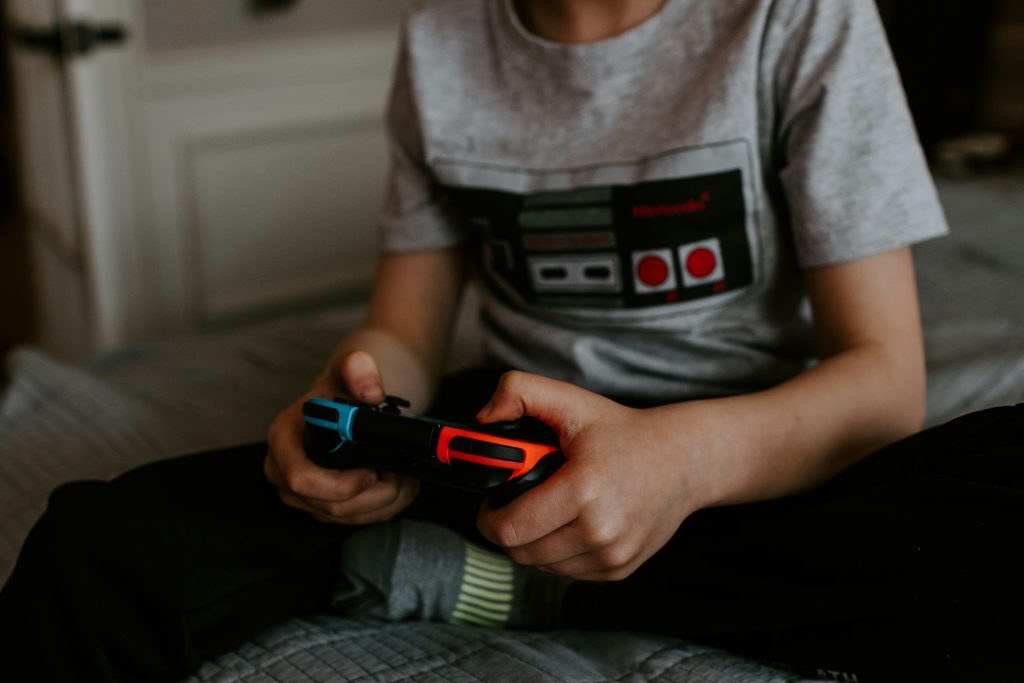 A child sits, holding a Nintendo Switch controller in their hands, playing a video game that's not in the frame. They're wearing a grey t-shirt that has a NES controller image on it.