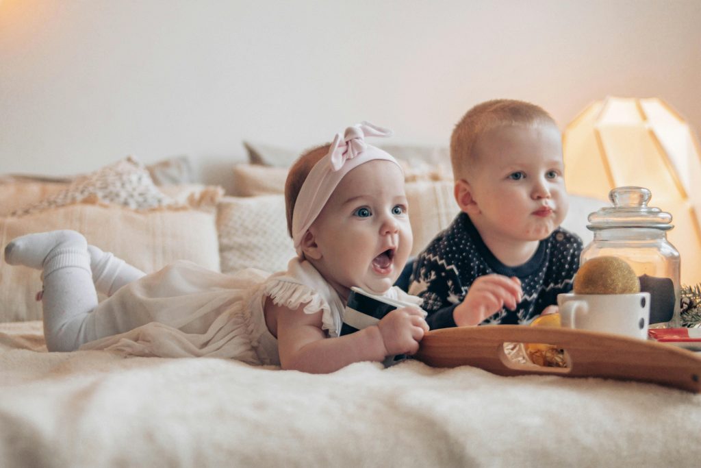 Two babies laid together on a bed, one dressed in pink frills with a bow and the other in a dark sweater.