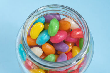 A colourless glass jar full of jellybeans in a range of colours: yellow, cyan, green, red, purple, orange, pink and white. The jar stands against a featureless blue background