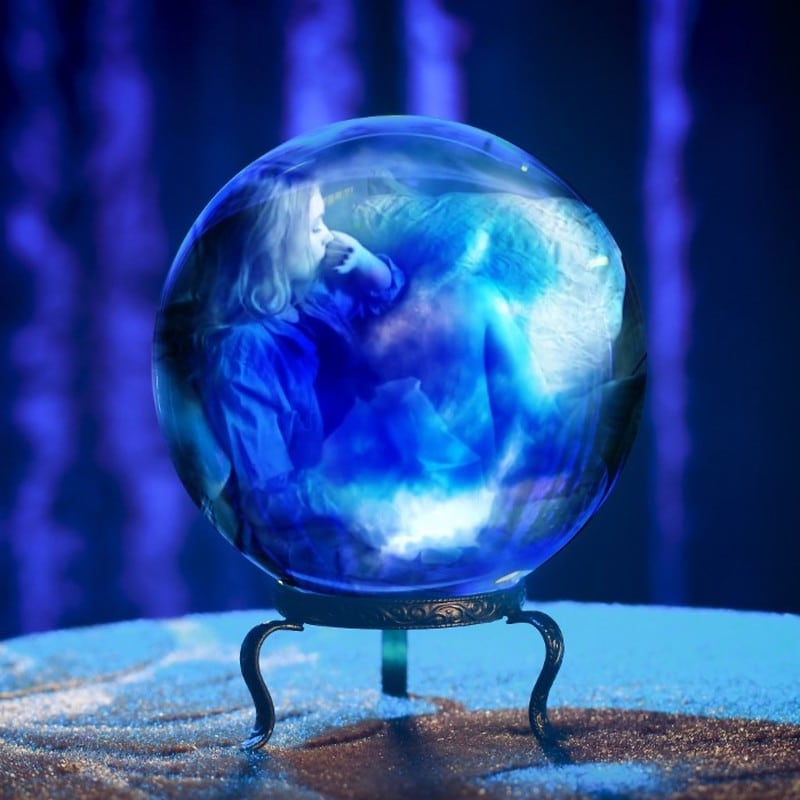 A crystal ball resting on a stand atop a table, with purple-blue curtains out of focus in the background. A dream-like image of two people, possibly a sleeping woman and man, shows inside the ball.