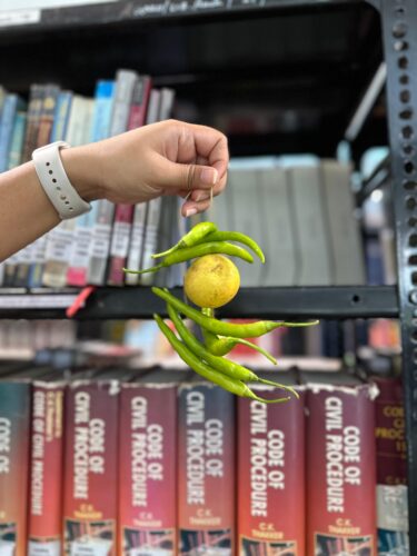 A hand wearing a white watch holds up a string of three green chillis, a lemon and four more green chillis, in front of some library shelves.