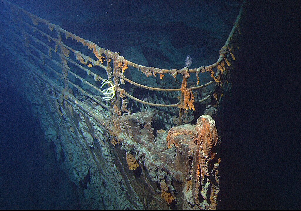 A photograph of the bow of the Titanic wreck in the Atlantic. The metal rails and hull are corroded, with assorted sea creatures growing on them and sitting around them.